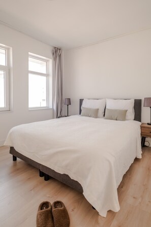 Une chambre avec un grand lit, deux tables de chevet, une fenêtre avec des rideaux et une paire de chaussons posés sur le sol.