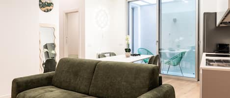 A modern living room with a green sofa, a dining area with a table and chairs, and a large sliding glass door leading to a balcony.