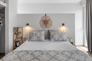 A bedroom with a large bed, two wall-mounted lamps, a decorative wall piece, and a shelf with items. - The Aer Element Of Gaea (Tinos)