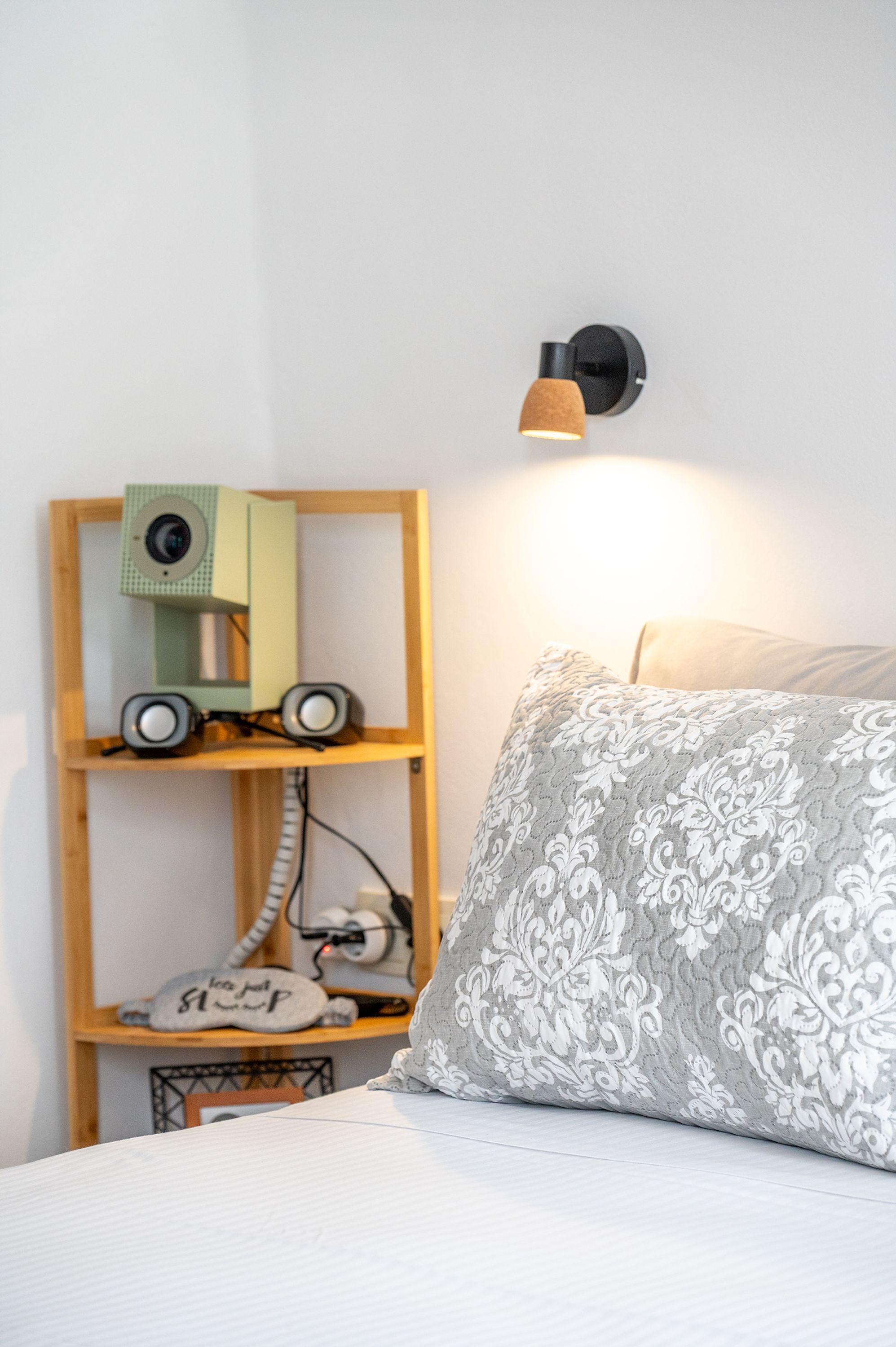 A bed with a patterned pillow, a shelf with electronic devices, and a wall-mounted light fixture.