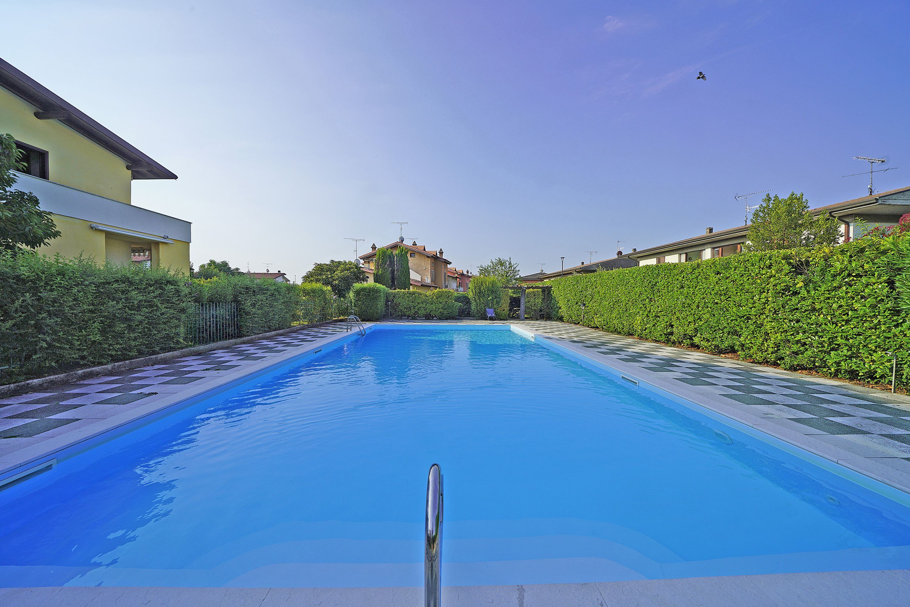 Uma piscina com superfície de água azul cristalina, rodeada por sebes bem aparadas e uma fileira de casas ao fundo.