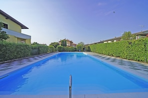 A swimming pool with a clear blue water surface, surrounded by neatly trimmed hedges and a row of houses in the background. - Raffa Holiday in Raffa (Puegnago del Garda)