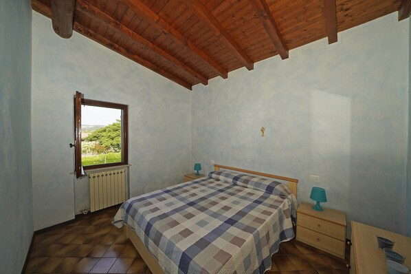Une chambre avec un plafond en bois, un lit recouvert d’une courtepointe à carreaux, une fenêtre donnant sur la verdure et un radiateur.
