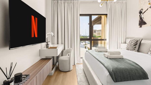 A modern hotel room with a bed, a desk, a chair, a TV, and a balcony with curtains. - Stayshort Stylish 1BR Apartment Near Burj Al Arab Family-friendly Sleeps 4 (Dubai)