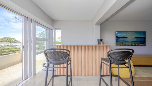 Una habitación moderna con un ventanal corredero que da a un balcón, dos sillas, un separador de madera con listones y un televisor en la pared, además de un otoman amarillo.