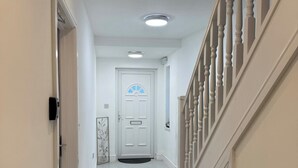 Un pasillo con una puerta blanca, un felpudo, una escalera con barandas blancas y una lámpara en el techo.