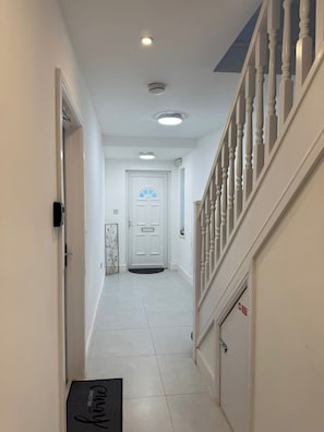 Un pasillo con una puerta blanca, un felpudo, una escalera con barandas blancas y una lámpara en el techo.