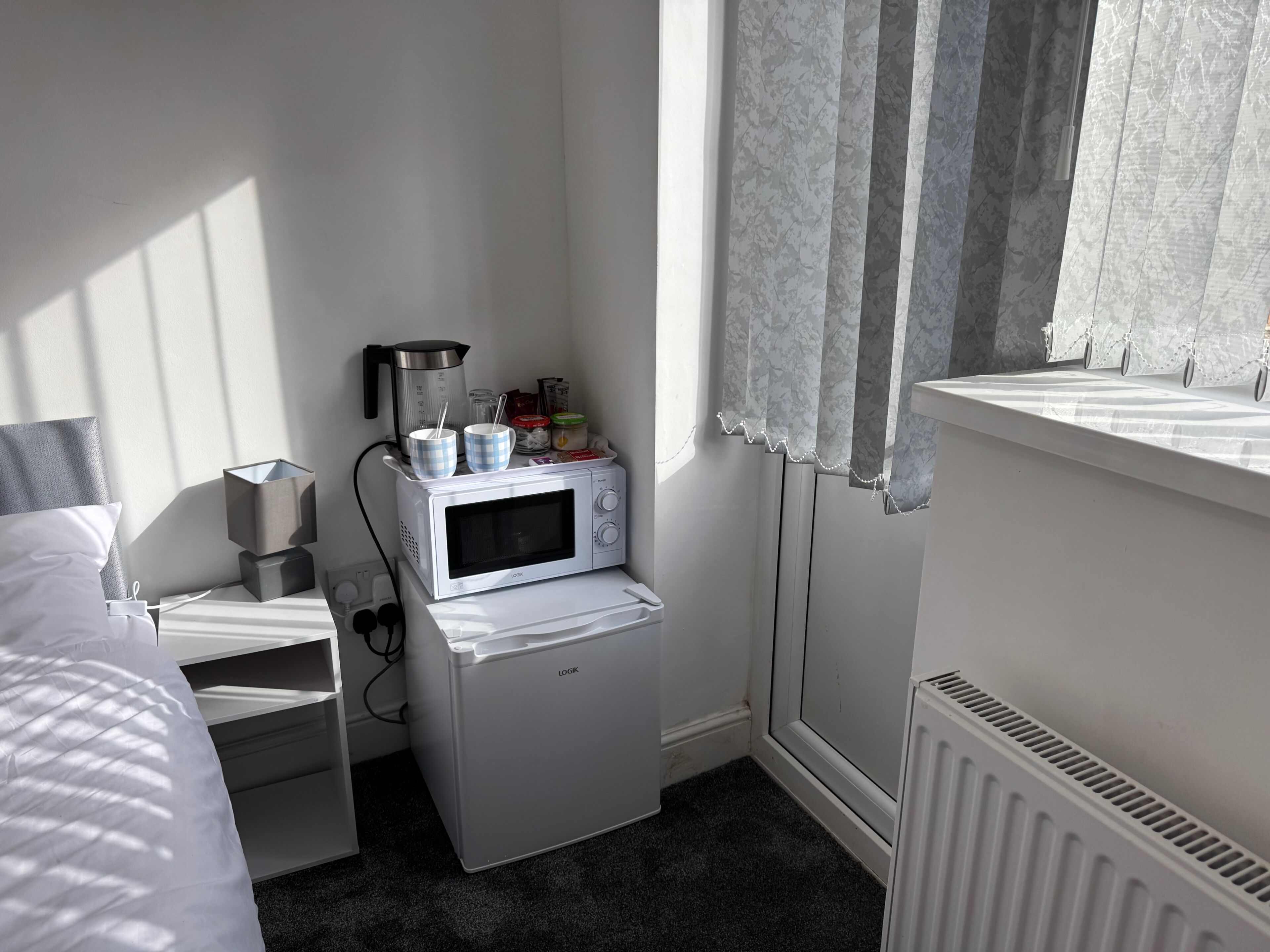 A compact hotel room with a bed, a small white cabinet, a microwave, and a radiator.