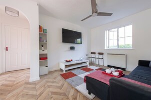 Una habitación moderna y luminosa con ventilador de techo, televisión de pantalla plana en la pared, un sofá blanco, un tapete rojo y gris, y una ventana que deja entrar luz natural.