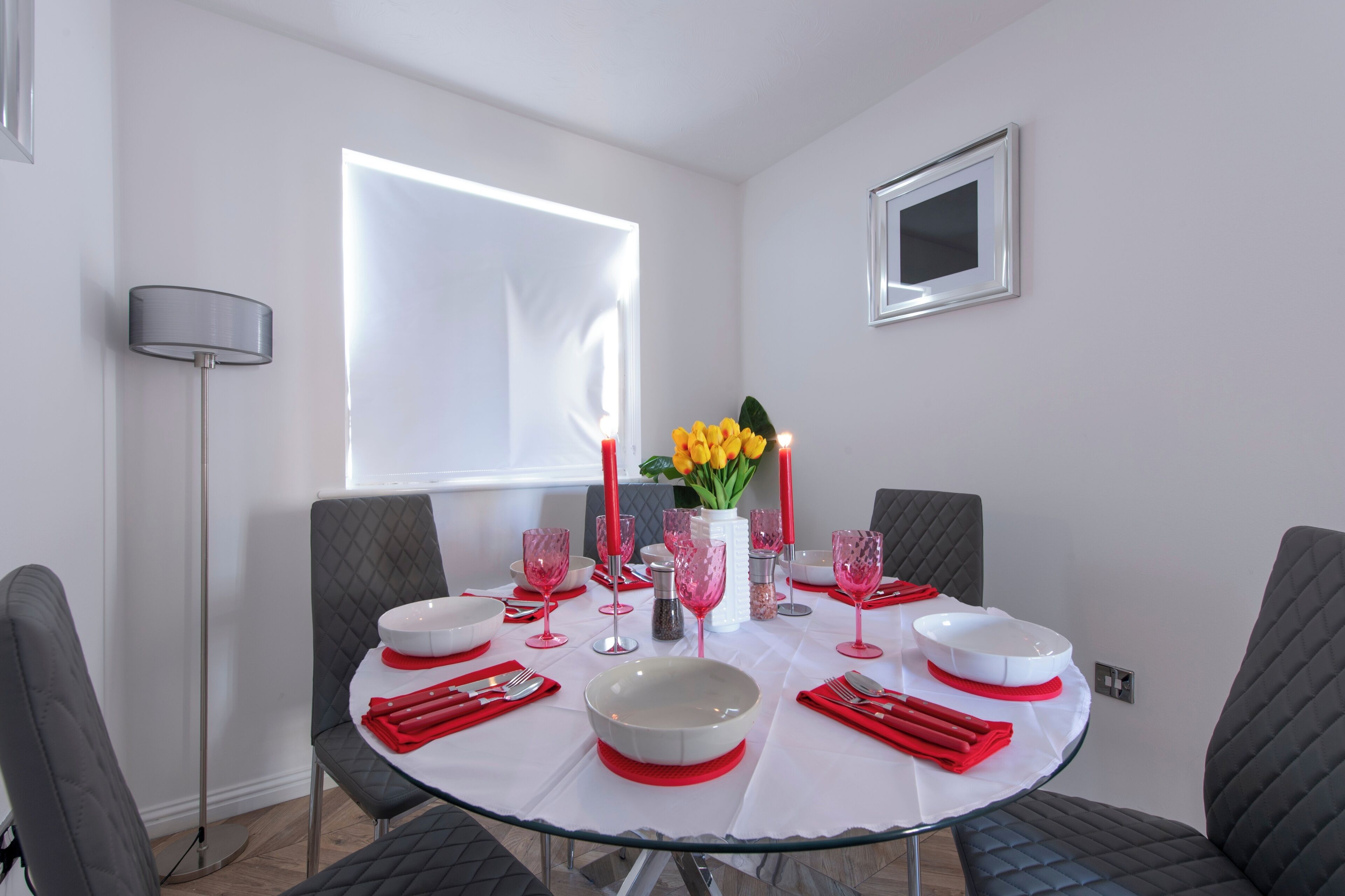 Un coin repas avec une table ronde dressée pour quatre personnes, agrémenté de vaisselle rouge et blanche, d’un vase avec des fleurs jaunes et d’une bougie rouge.