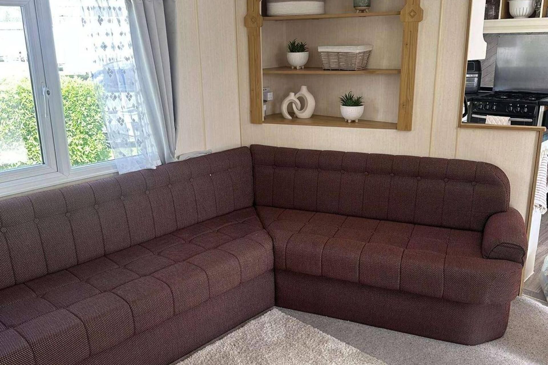 A corner living room with a dark brown sectional sofa, a wooden shelf with decorative items, and a kitchen area with a stove.