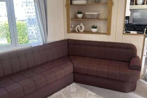 A corner living room with a dark brown sectional sofa, a wooden shelf with decorative items, and a kitchen area with a stove. - 8 Berth Caravan at Butlins Skegness (Skegness)
