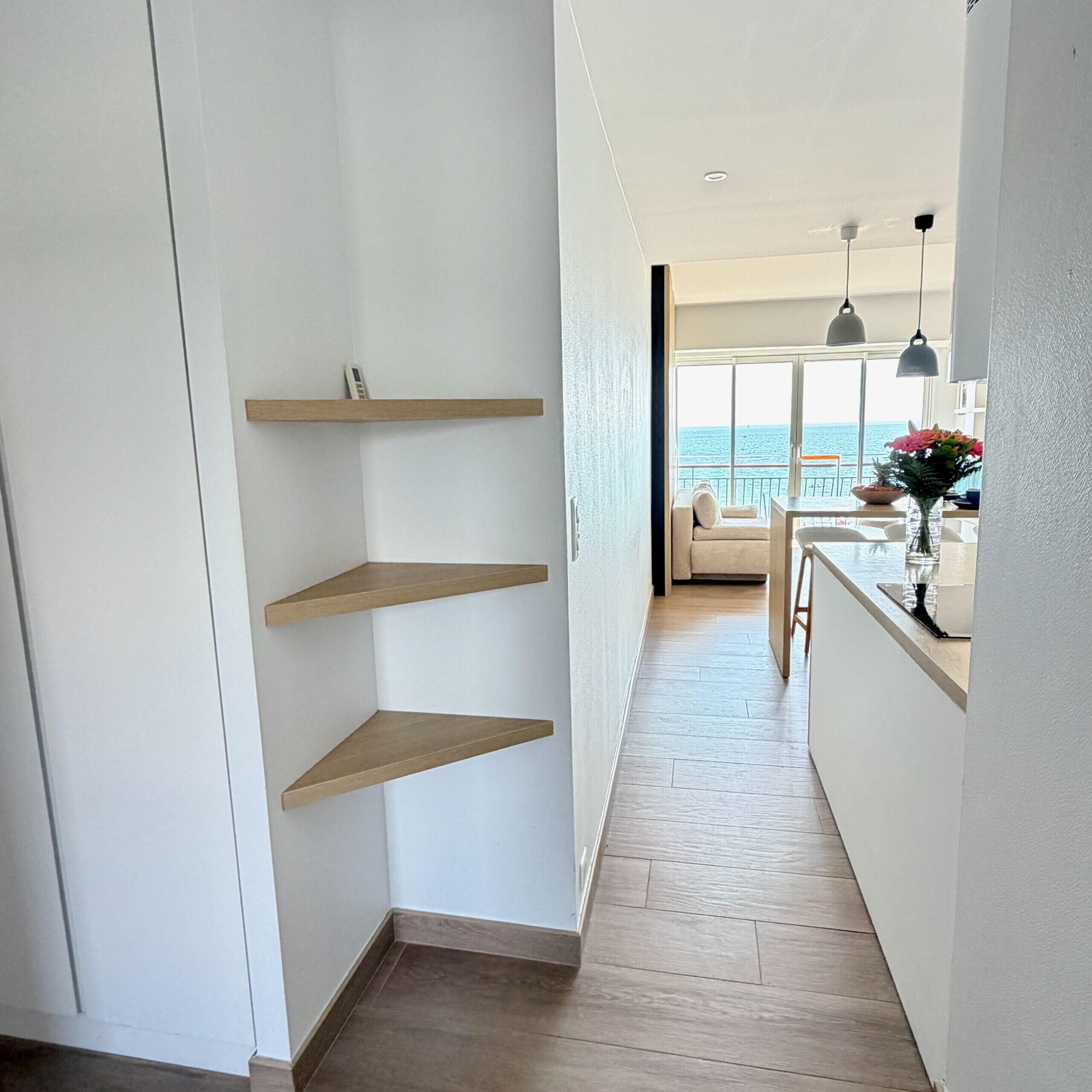 A corridor with wooden shelves, a view to a balcony with a sofa and table, and a wooden floor.
