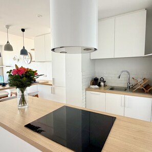 A modern kitchen with white cabinets, a black induction cooktop, a stainless steel sink, and pendant lighting. - Face mer à La Baule : Evobaule (La Baule-Escoublac)