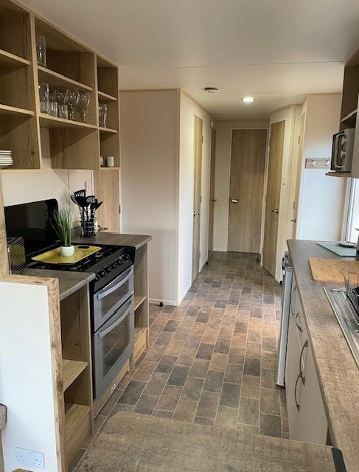 A narrow kitchen with a built-in oven, a stove, and a wooden shelf with glassware.