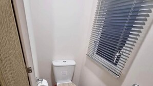 A small bathroom with a toilet, a bidet, a toilet paper holder, and a window with blinds. - 8 Berth Caravan Butlins Skegness (Skegness)