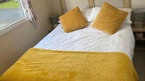 A single bed with a wooden headboard, two pillows, a bedside table with a lamp, and a window with curtains. - 8 Berth Caravan Butlins Skegness (Skegness)