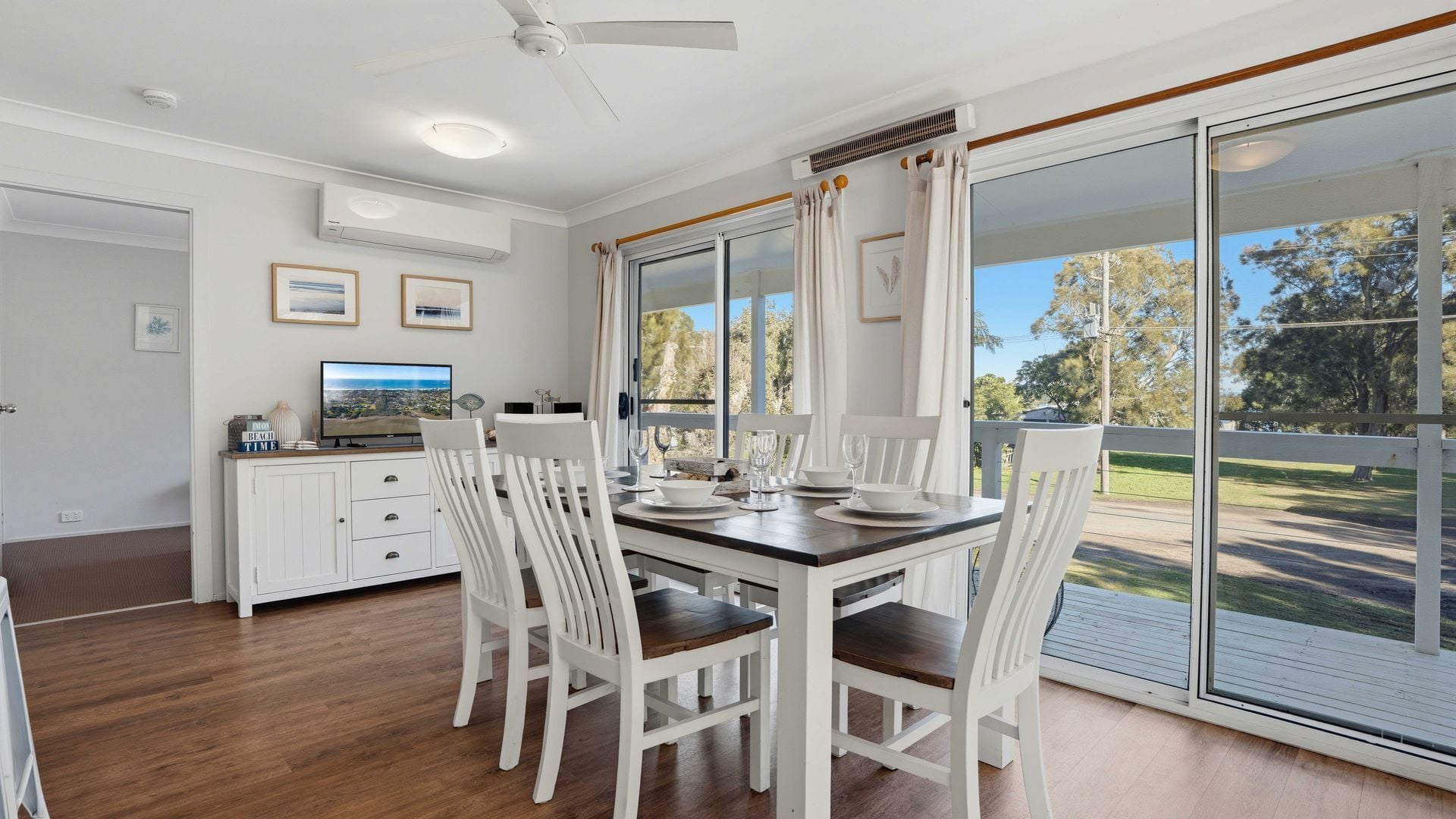 A dining area with a table set for four, white chairs, a television on a cabinet, and a view through large glass doors.