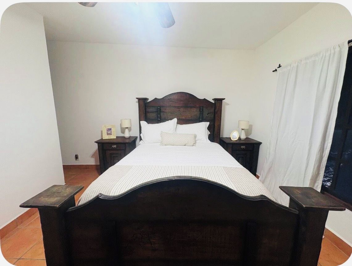 A bedroom with a wooden bed, white bedding, two bedside tables with lamps, and a window with curtains.