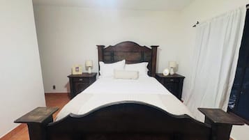 A bedroom with a wooden bed, white bedding, two bedside tables with lamps, and a window with curtains.
