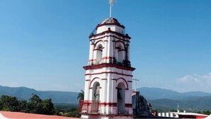 A white and red bell tower with a cross on top, surrounded by a clear blue sky and distant mountains.
