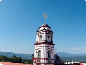 A white and red bell tower with a cross on top, surrounded by a clear blue sky and distant mountains.