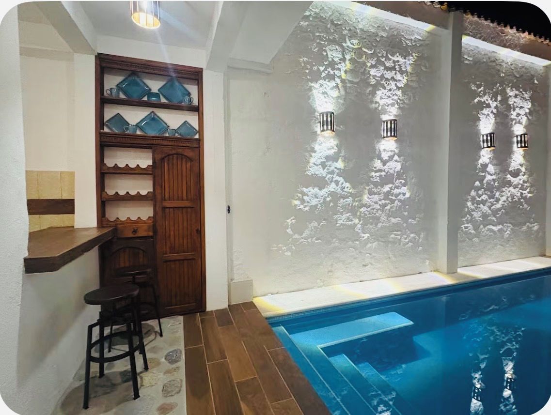 Zona de piscina con bar y taburetes, repisa de madera con objetos azules y luces empotradas en la pared.