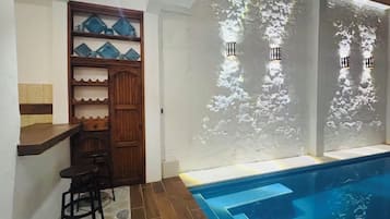 Zona de piscina con bar y taburetes, repisa de madera con objetos azules y luces empotradas en la pared.