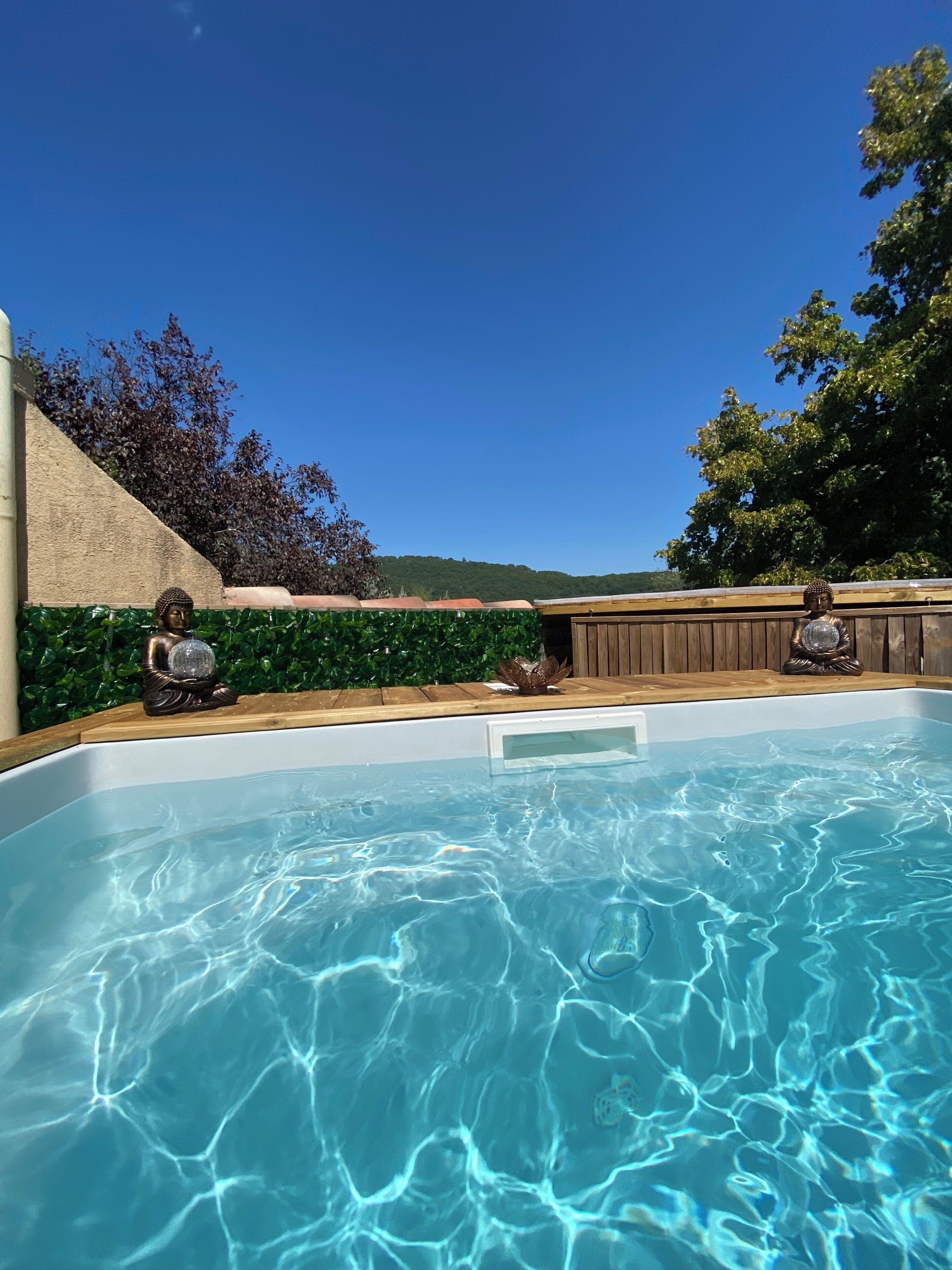 Une piscine ornée de deux statues de Bouddha, entourée d’une terrasse en bois et d’une haie verdoyante.