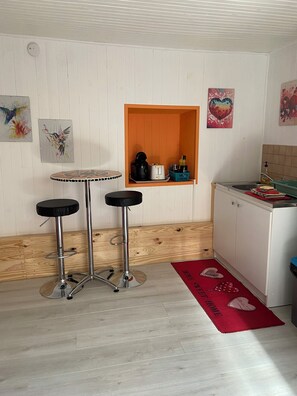 Eine kleine Küche mit Holzarbeitsplatte, Spüle und einem orangen Aufbewahrungsfach. Zwei Hocker und eine rote Fußmatte mit dem Schriftzug 'HOME'.