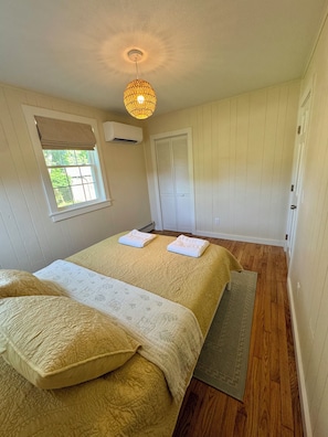 A bedroom with a bed, a window, an air conditioning unit, a door, and a ceiling light fixture.