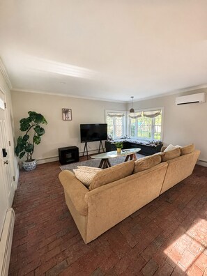 A living room with a beige sofa, a TV on a stand, a potted plant, and a window with curtains.