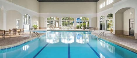 Une grande piscine intérieure à l’eau d’un bleu limpide, entourée d’espaces aménagés pour s’asseoir et dotée de grandes fenêtres.