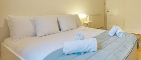 Cama bem arrumada com roupa de cama branca e um edredom azul, uma porta branca e uma mesa de cabeceira com abajur.
