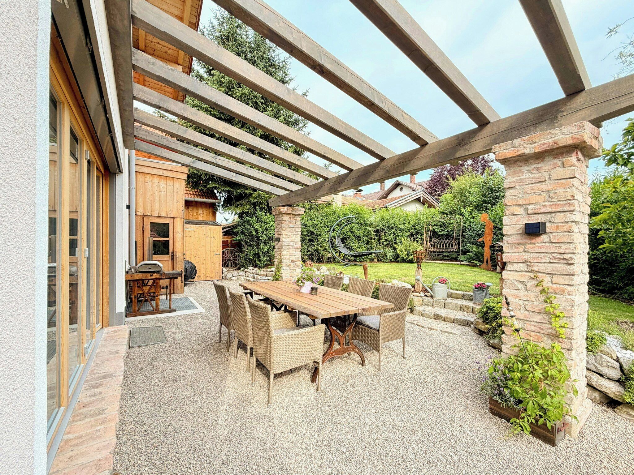 An outdoor patio area with a wooden pergola, a dining table with chairs, and a stone wall.
