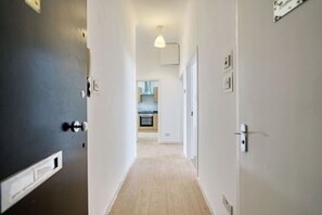 Um corredor com uma porta à esquerda, uma luminária acima e uma cozinha visível ao longe.