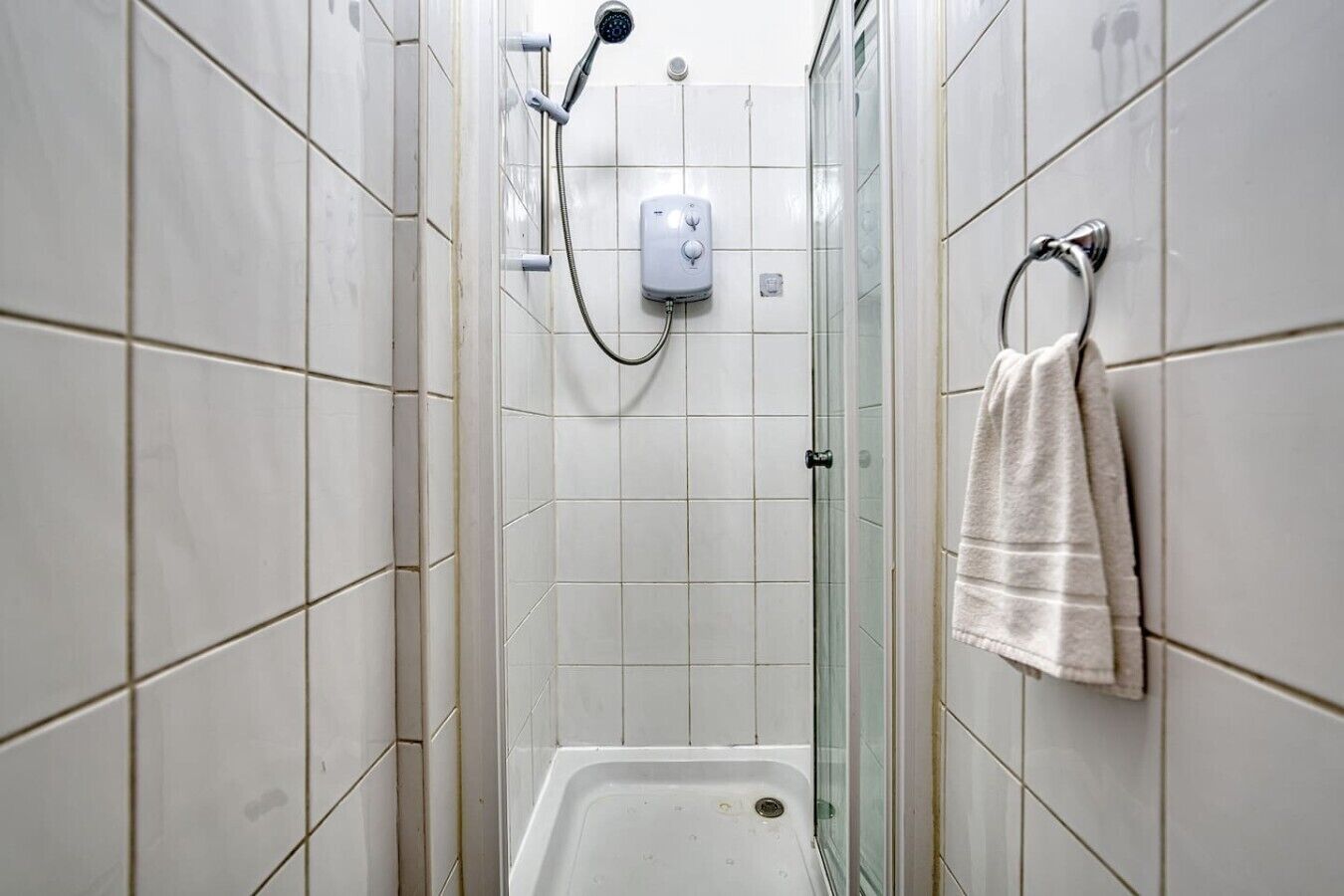 A narrow shower stall with white tiles, a handheld showerhead, and a towel rack with a towel.