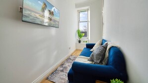 Een woonkamer met een blauwe bank, een flatscreen televisie aan de muur, een raam met uitzicht en een kleine plant in een pot op het raamkozijn.