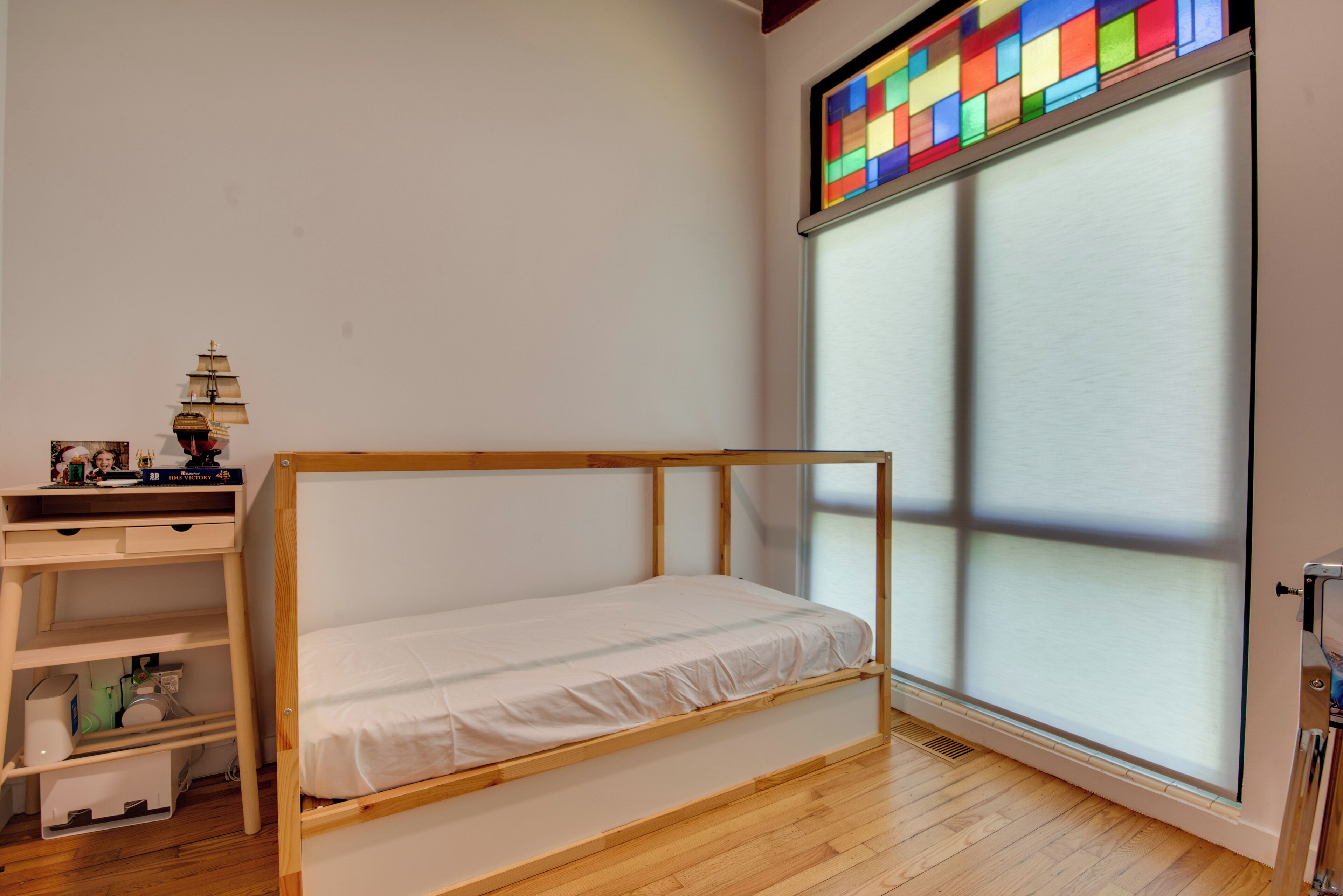 A room with a wooden bunk bed, a desk with a lamp, and a large window with a colorful stained glass design.