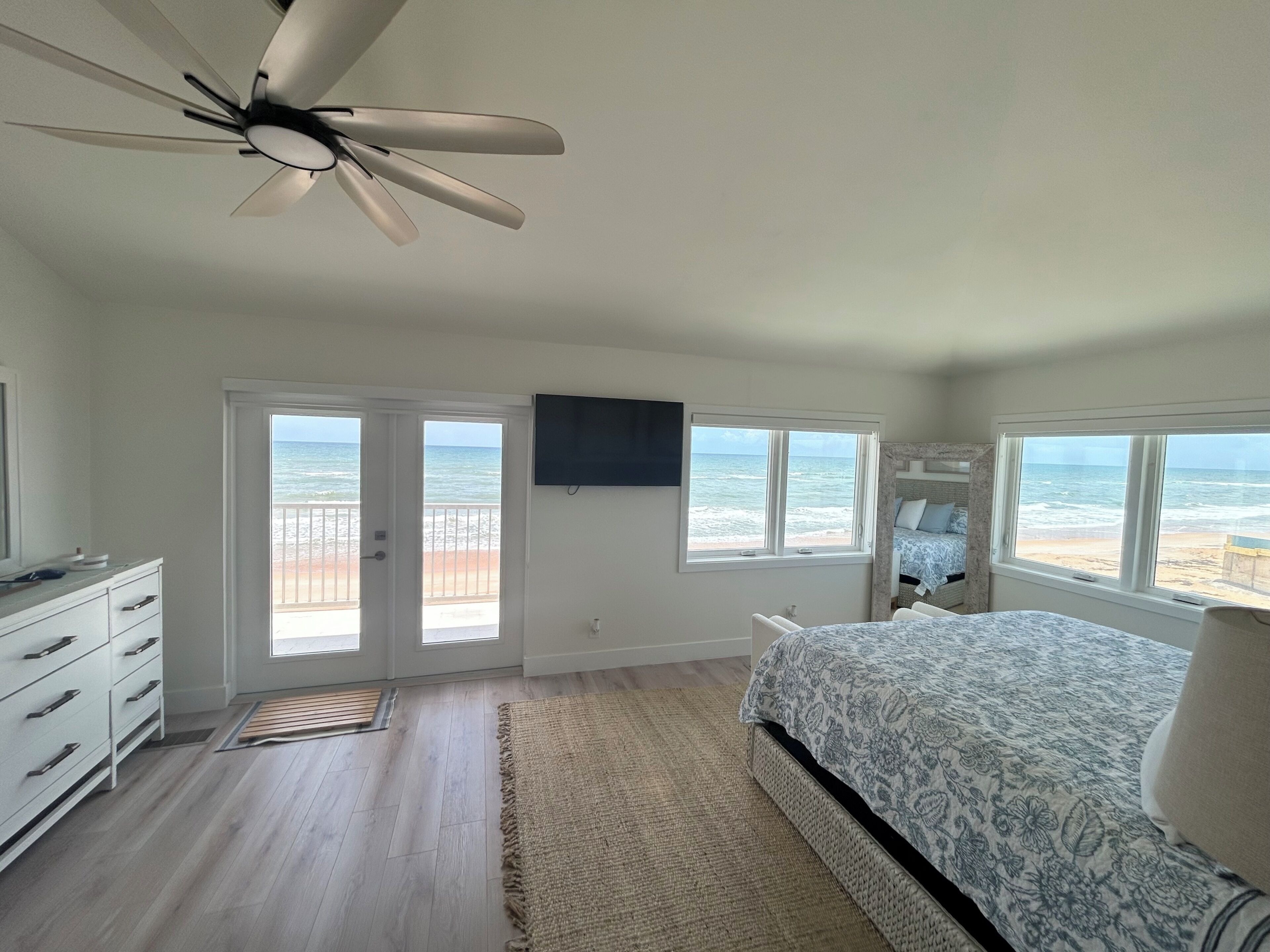 A bedroom with a bed, a ceiling fan, a TV, and a view of the beach.