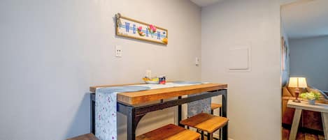 Una pequeña cocina con una mesa de madera y taburetes de bar, un interruptor de luz en la pared y un cartel enmarcado sobre la encimera.