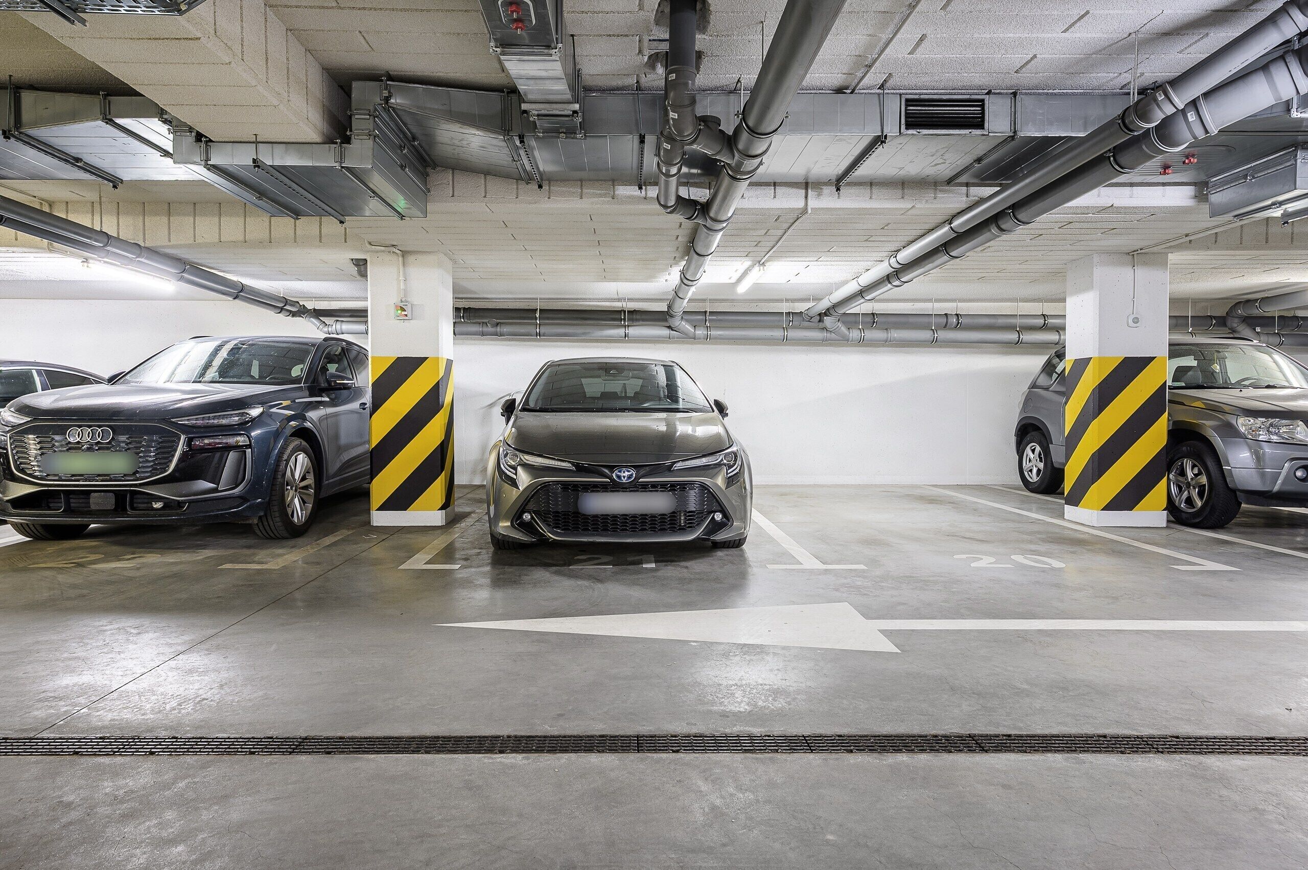 A parking garage with three cars parked in designated spaces, concrete pillars with yellow and black stripes, and directional arrows on the floor.
