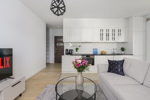 A modern living room with a sofa, a coffee table with a vase of flowers, a TV, and a kitchen area with cabinets and appliances. - Lumina modern Apart parking 2 balcony (Warszawa)