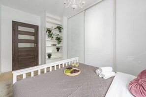 A bedroom with a bed, a bedside table with a fruit platter, a white railing, a wooden door, and a white shelving unit with plants. - Lumina modern Apart parking 2 balcony (Warszawa)