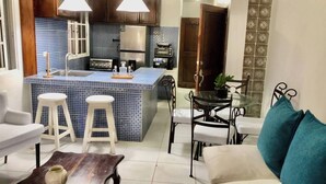 Un ambiente compatto con angolo cottura, zona pranzo con tavolo in vetro e salotto con divano e due sgabelli.