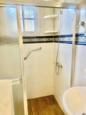 Ein Badezimmer mit Duschkabine aus Glas, Holzboden, weißem Waschbecken und einem Fenster mit Jalousien.