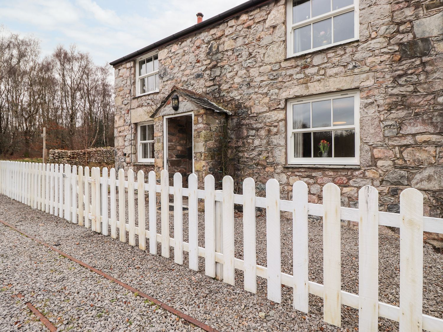 A stone house with a white picket fence, a stone wall, and a gravel driveway.