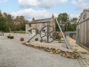 A rustic stone building with a white fence, a large metal gear sculpture, and a gravel path. - Irt Cottage (Seascale)