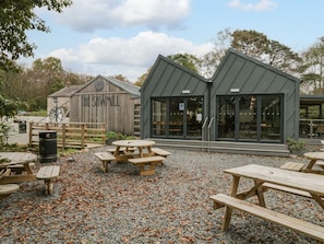 An outdoor seating area with wooden picnic tables and a gravel ground, surrounded by a wooden fence and a building labeled 'The Sawmill'. - Irt Cottage (Seascale)