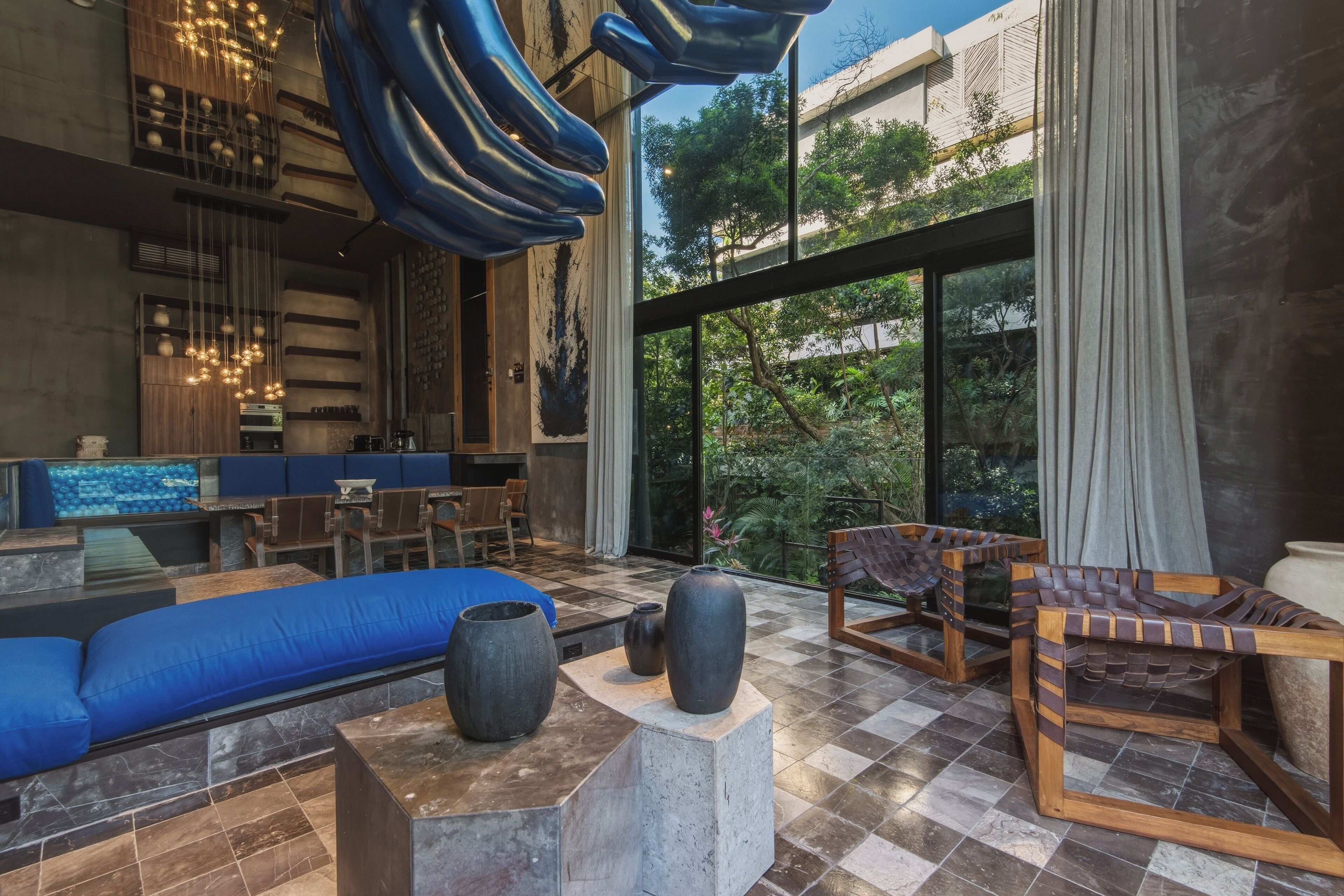 A modern interior with a blue sofa, wooden chairs, a large vase, and a view of greenery outside.